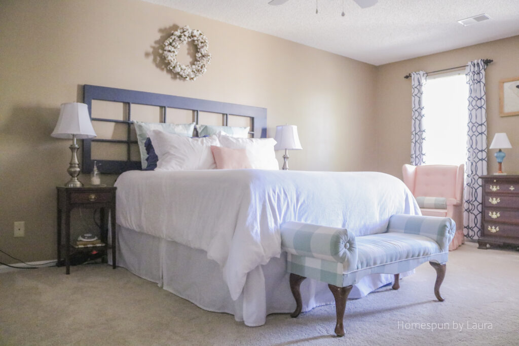 Master bedroom with white linens and navy blue antique french door headboard; mint gingham upholstered bench; pink chair